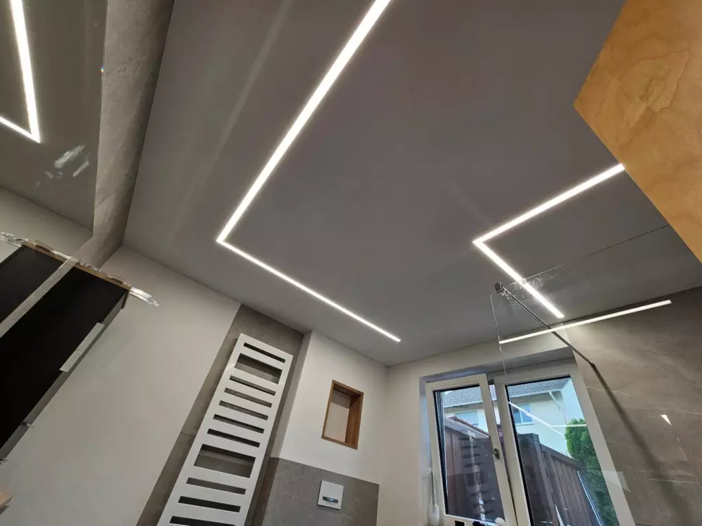 Bad mit heller Textilspanndecke und LED-Lichtlinien im 90°-Winkel, darunter Handtuchheizkörper, Fenster und Dusche