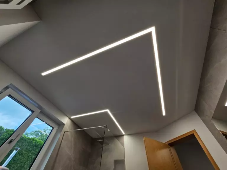 Spanndecke im Bad mit zwei winkelförmigen LED-Lichtlinien über Fenster und Glasdusche