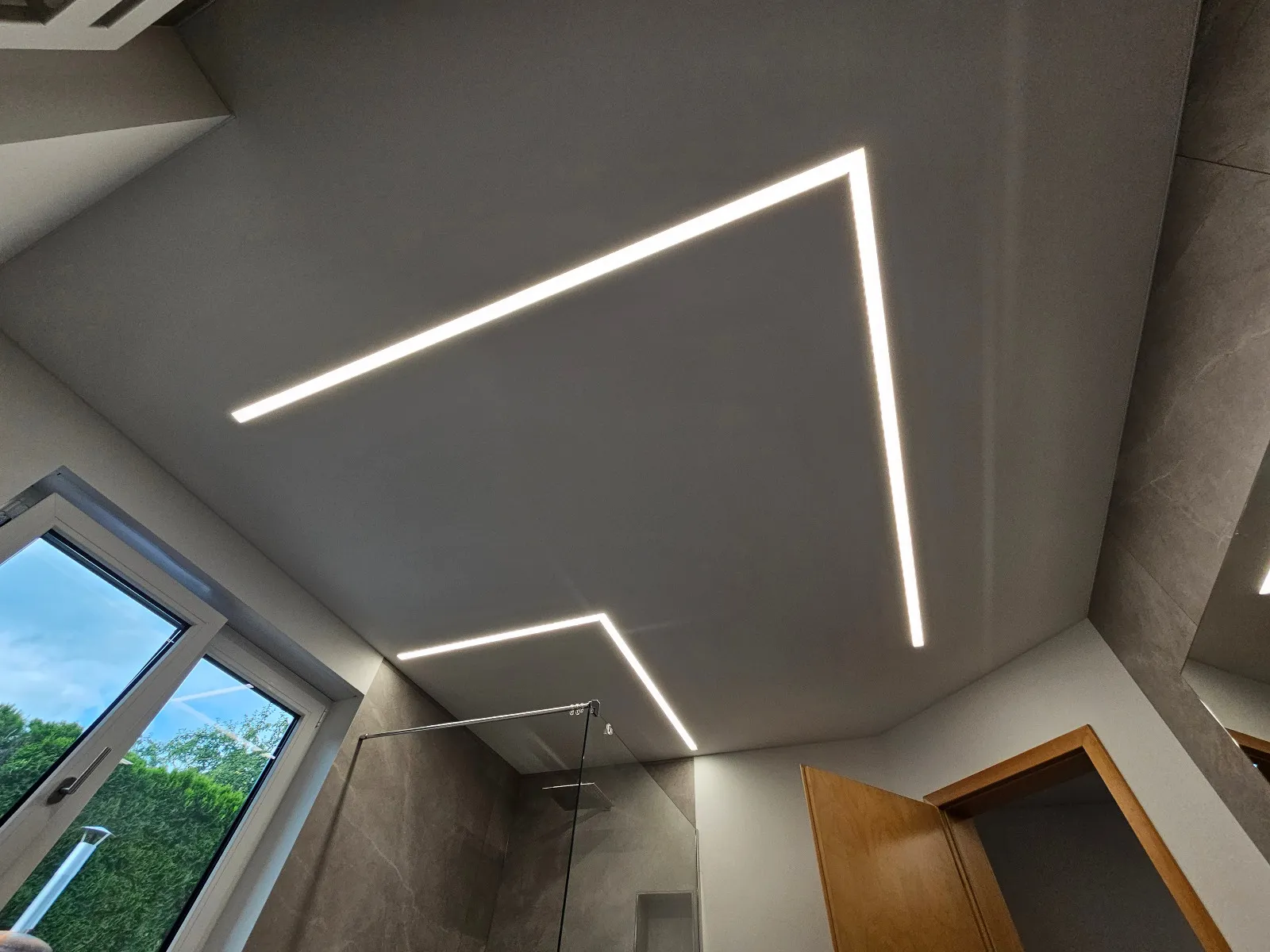Spanndecke im Bad mit zwei winkelförmigen LED-Lichtlinien über Fenster und Glasdusche