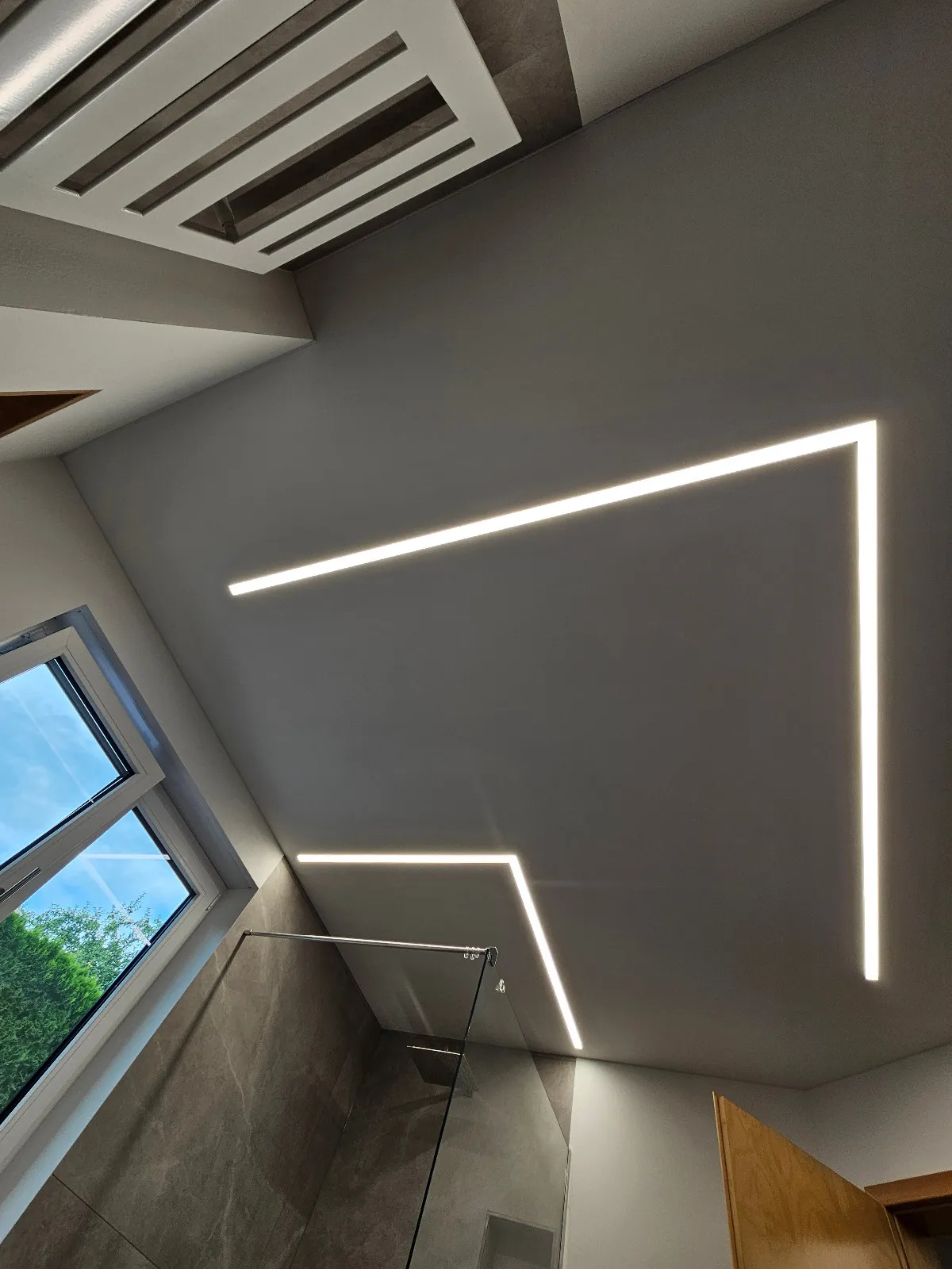 Blick nach oben: LED-Lichtlinien bilden zwei rechte Winkel in der Spanndecke über der Dusche