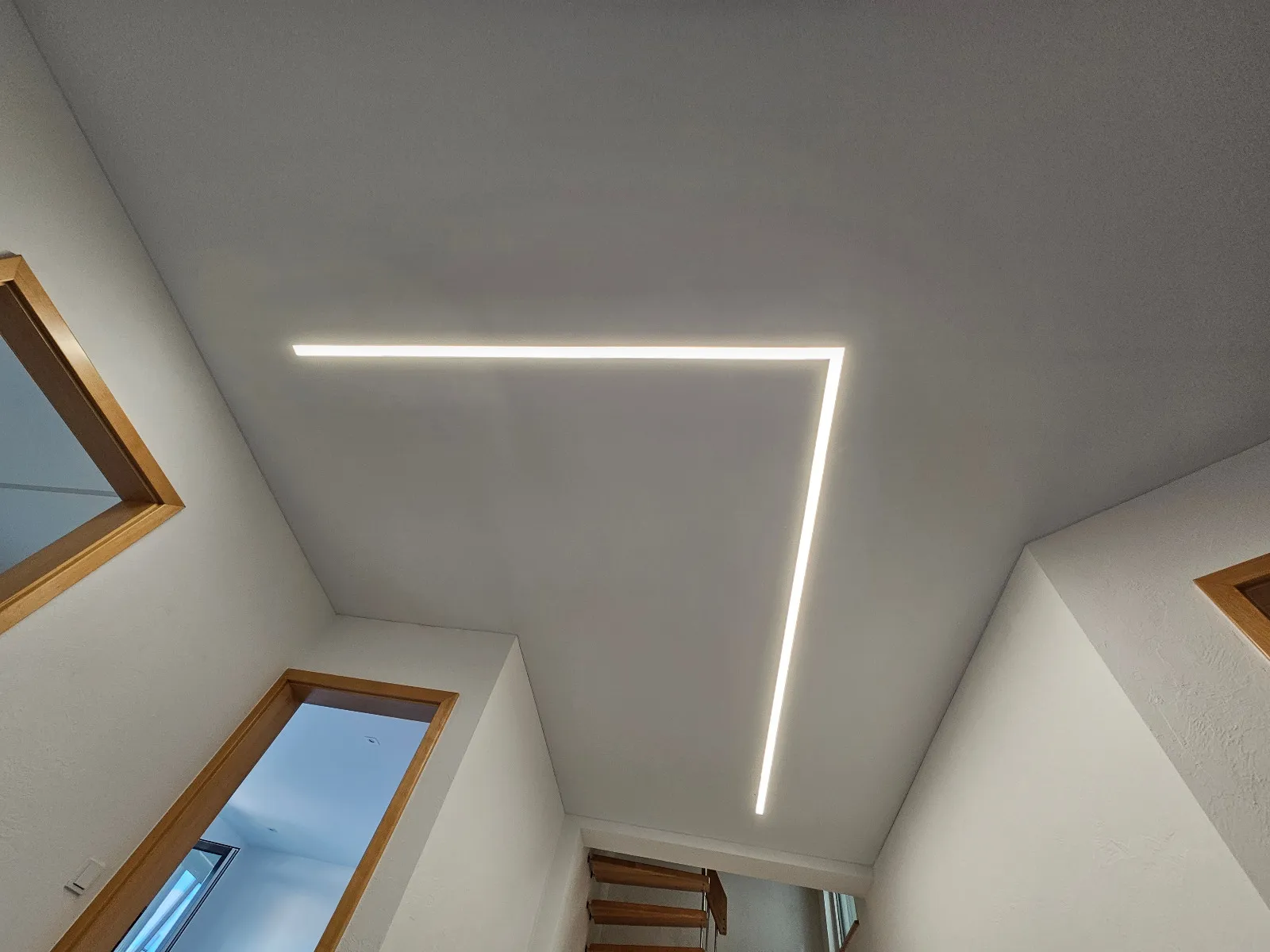 Flur mit Spanndecke und einer LED-Lichtlinie im 90°-Winkel, im Hintergrund eine Holztreppe
