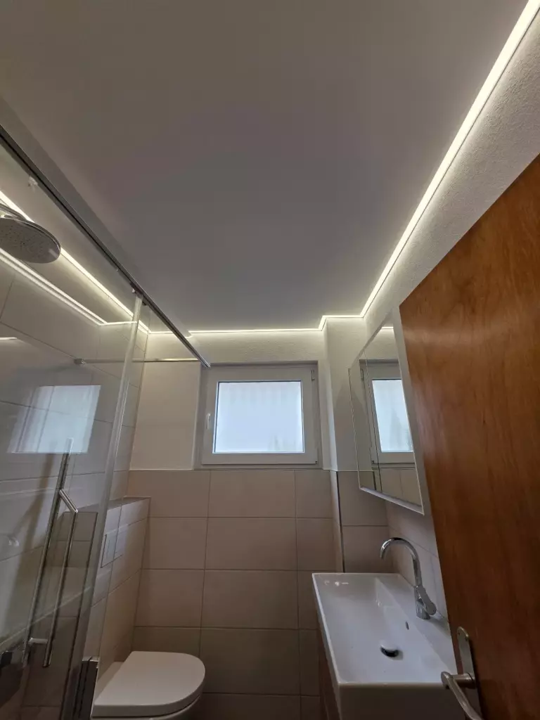 Nachher: Bad mit glatter weißer Spanndecke und umlaufender indirekter Randbeleuchtung, Fenster, Dusche und Waschbecken